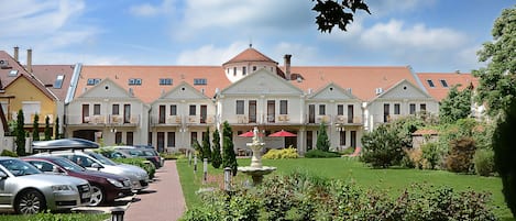 Un hotel con techo de tejas rojas, un patio central con fuente y una fila de coches estacionados.