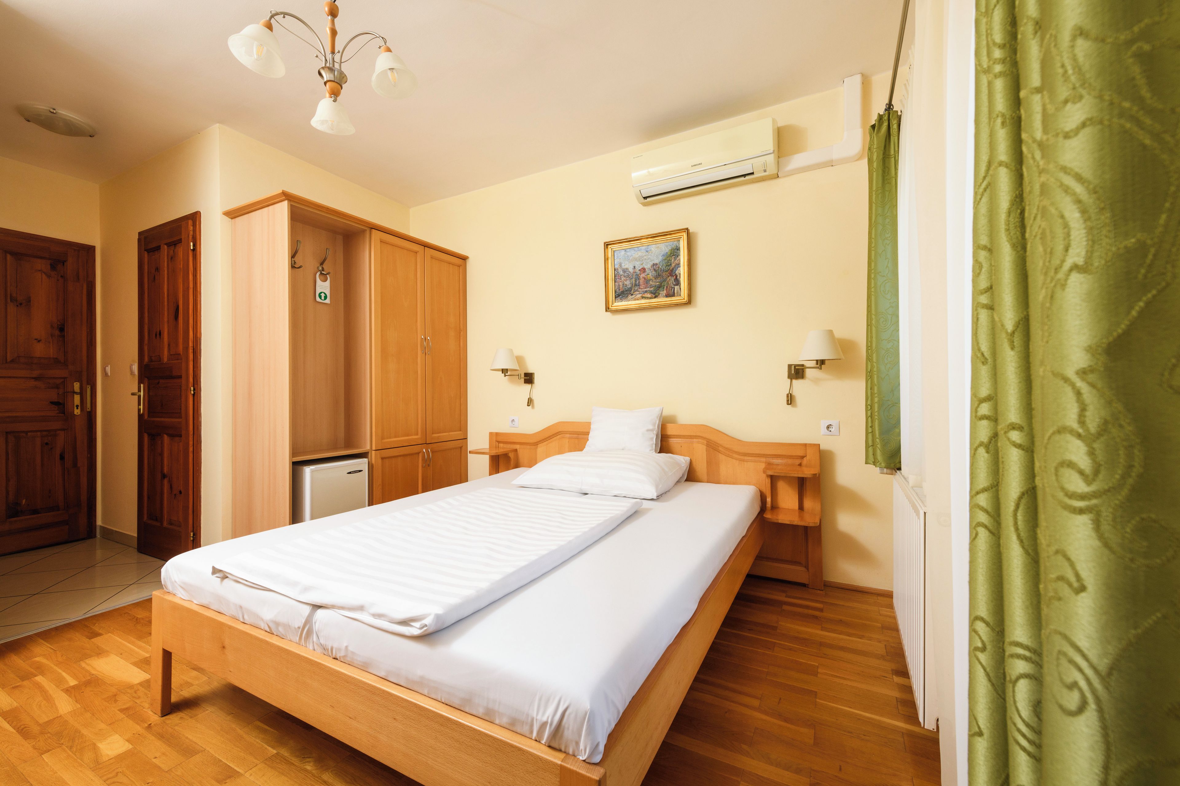 Habitación de hotel con una cama de madera, un armario, una mesita y aire acondicionado, además de un cuadro colgado en la pared.