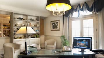 A hotel room with a desk, chair, lamp, shelves with various items, a potted plant, and a television.