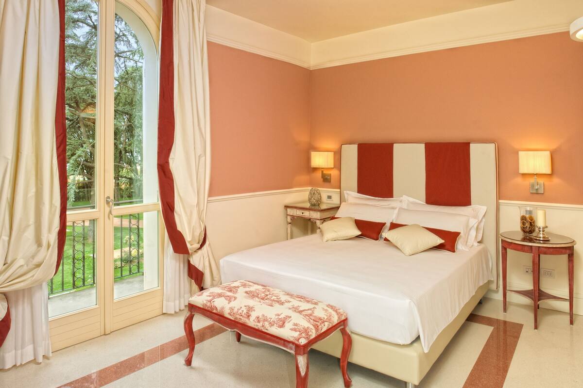 Une chambre à coucher avec un grand lit, un banc, deux tables de chevet et une vue sur un jardin, visible à travers une porte vitrée.