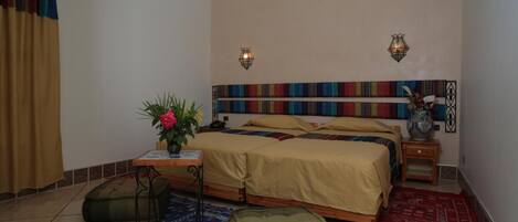 Un dormitorio con una cama, dos mesitas de noche, una mesita auxiliar, una pequeña mesa con un florero y un cabecero colorido.