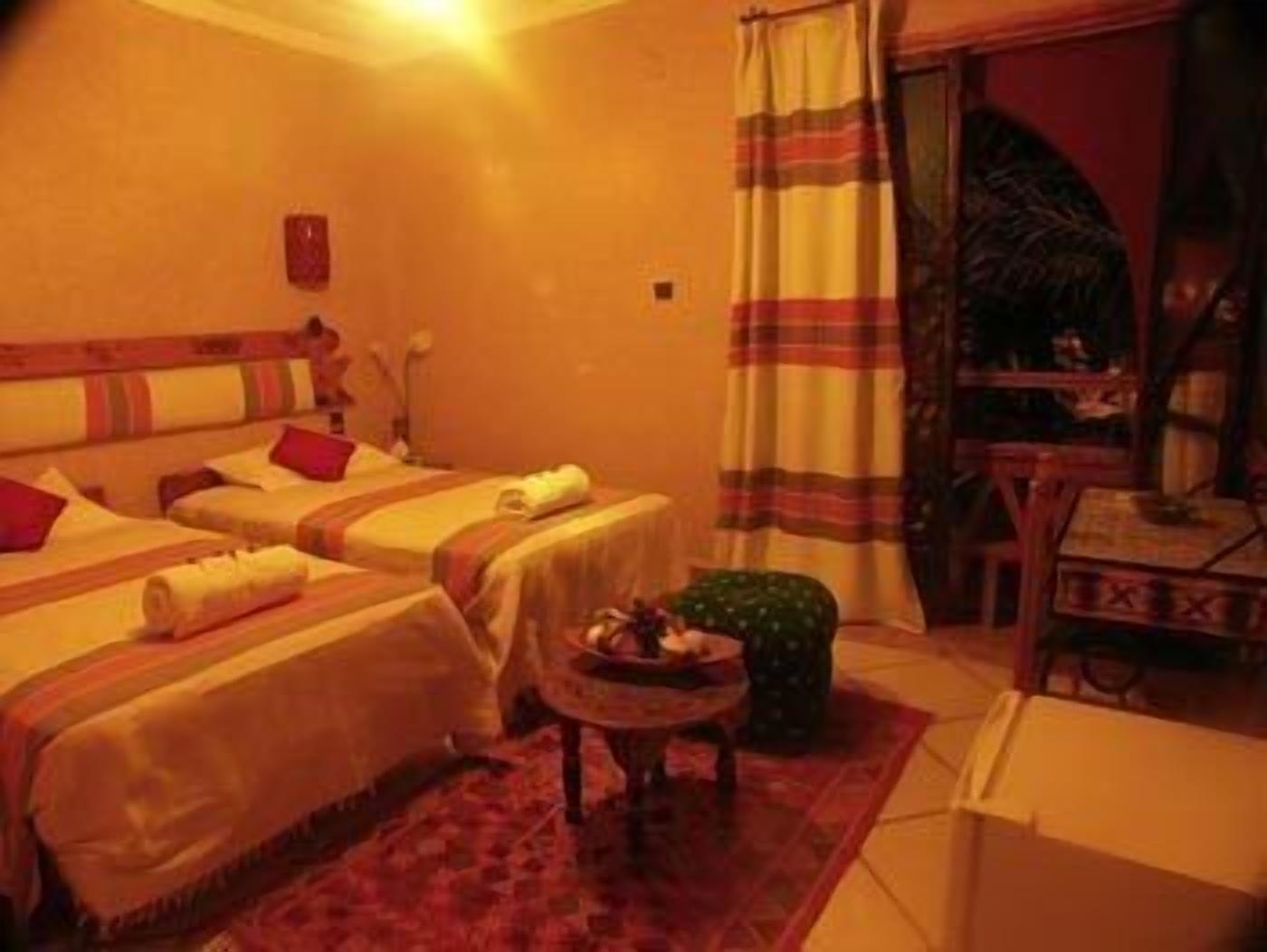 A hotel room with two beds, a small table, a chair, and a window with curtains.