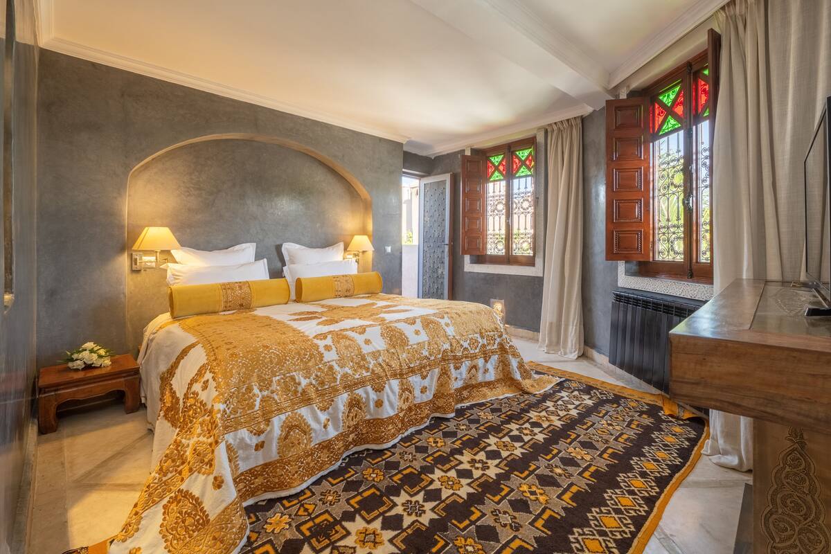 Quarto com uma cama grande, mesa de cabeceira, cabeceira de madeira, tapete estampado e janela com vidro decorativo.
