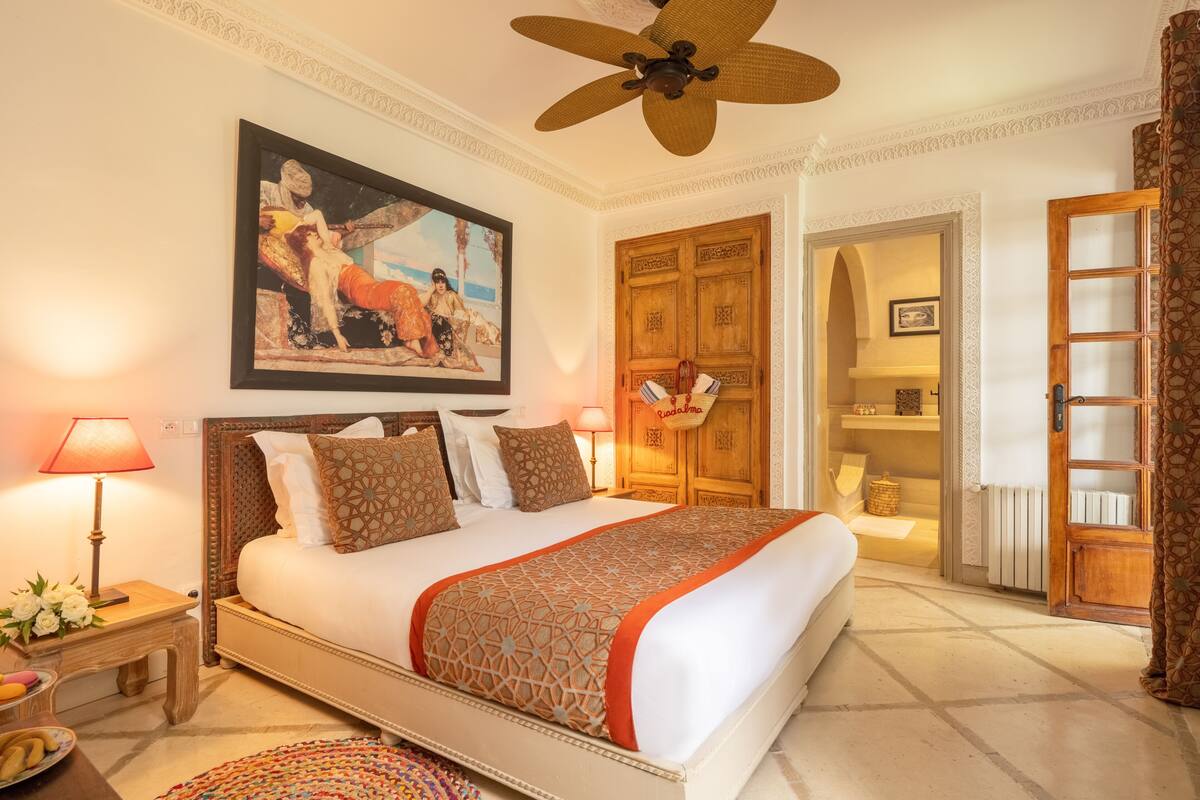 Um quarto espaçoso com uma cama grande, um ventilador de teto, um guarda-roupa de madeira e um quadro na parede.