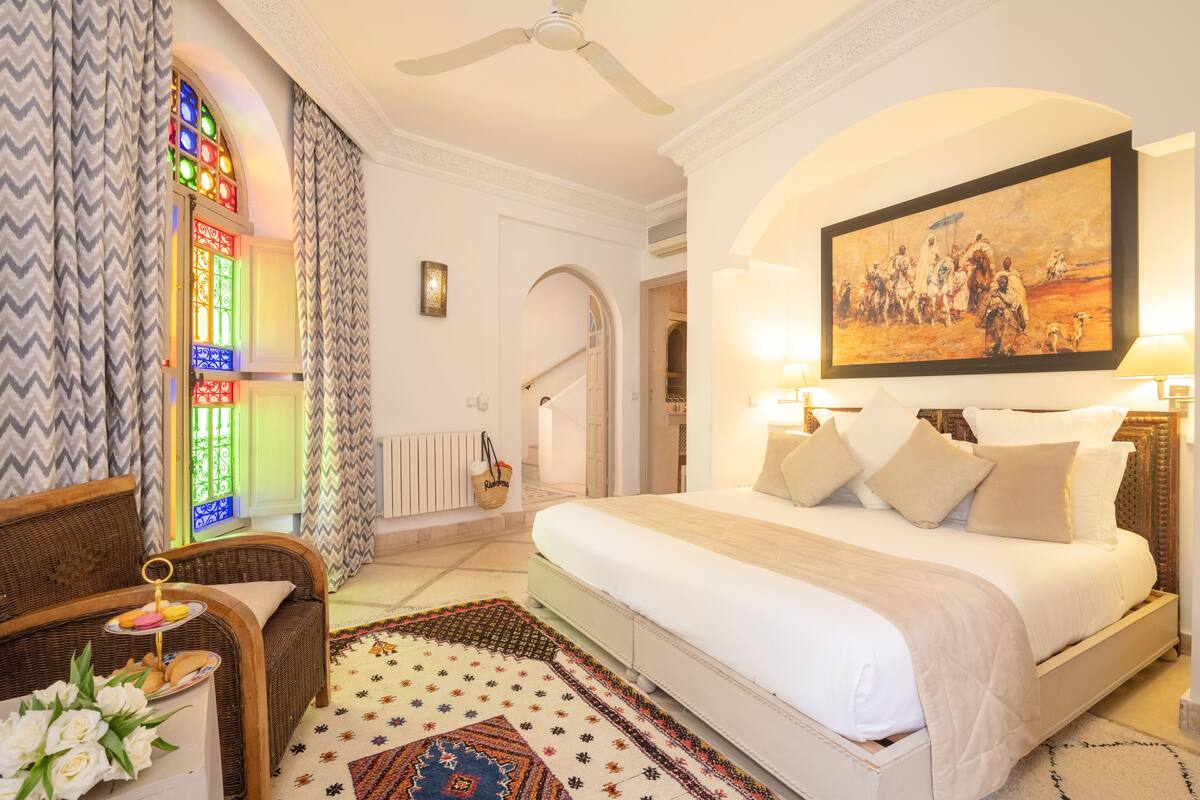 Quarto de hotel com uma cama grande, uma cadeira de vime, um tapete colorido e uma janela de vitrais.
