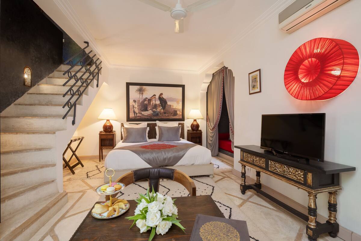 Quarto de hotel com cama, televisão, mesa de jantar com comida, escada e um objeto decorativo vermelho na parede.