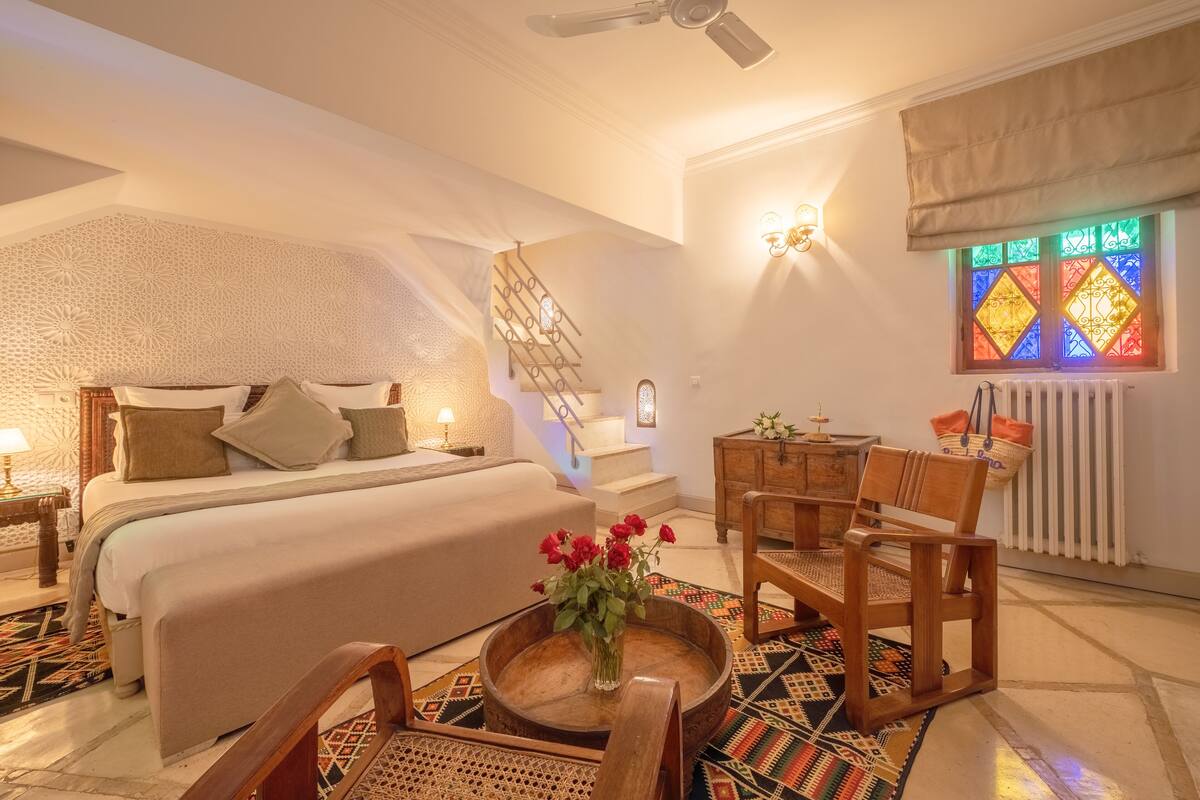 Um quarto com uma cama, uma cadeira de madeira, uma mesa pequena com um vaso de flores, uma escada e uma janela de vitrais.