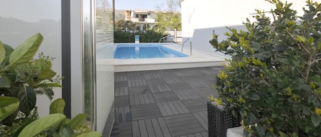 Uma área de piscina moderna ao ar livre, com um deck de madeira, cercada por vegetação e uma parede branca.
