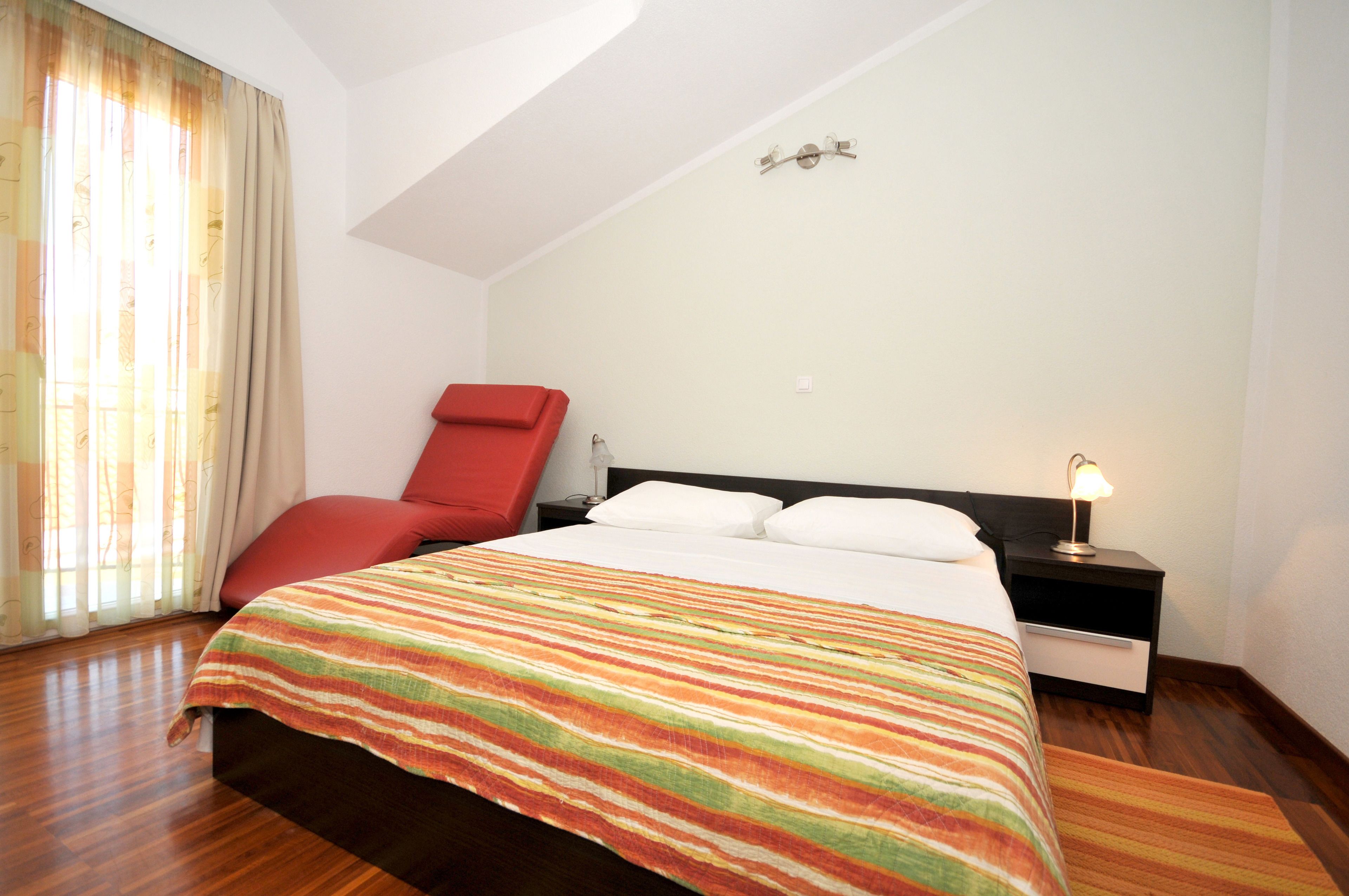A bedroom with a bed, a red chair, a nightstand with a lamp, and a window with curtains.