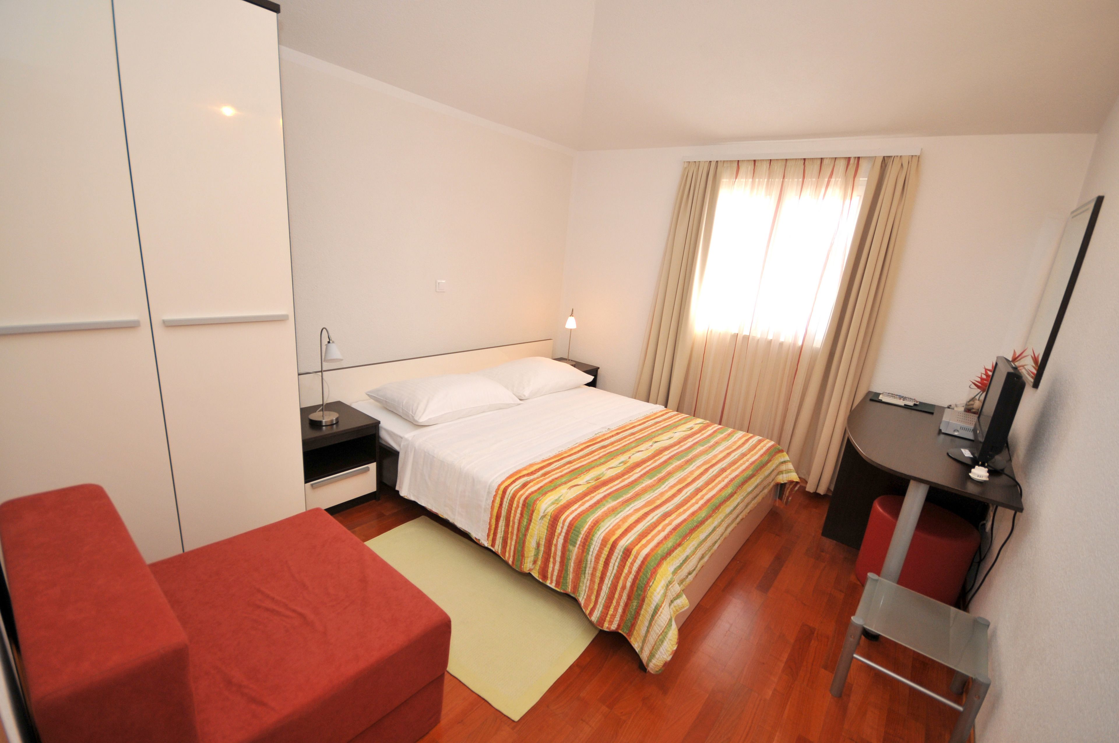 A hotel room with a bed, a desk, a chair, a red sofa, and a window with curtains.