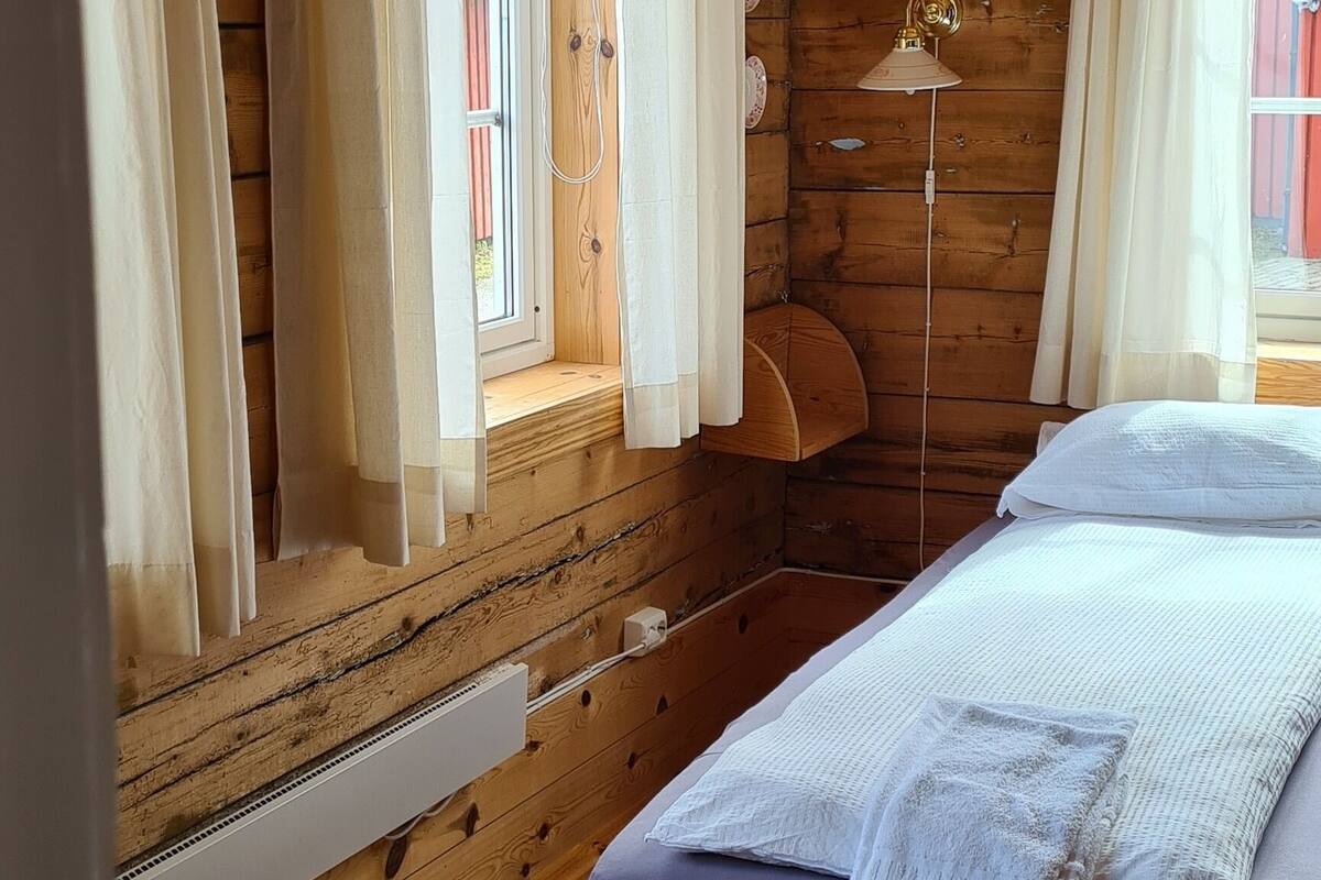 Una habitación de cabaña de madera con cama, cortinas, una lámpara y una ventana.