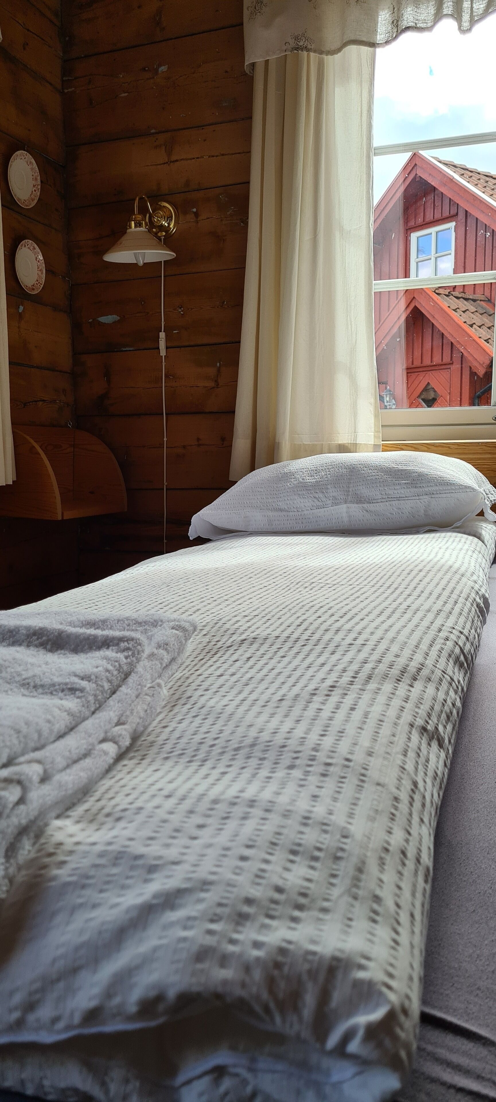 Une pièce de cabane en bois comprenant un lit, une lampe et donnant sur un bâtiment rouge situé à l’extérieur.