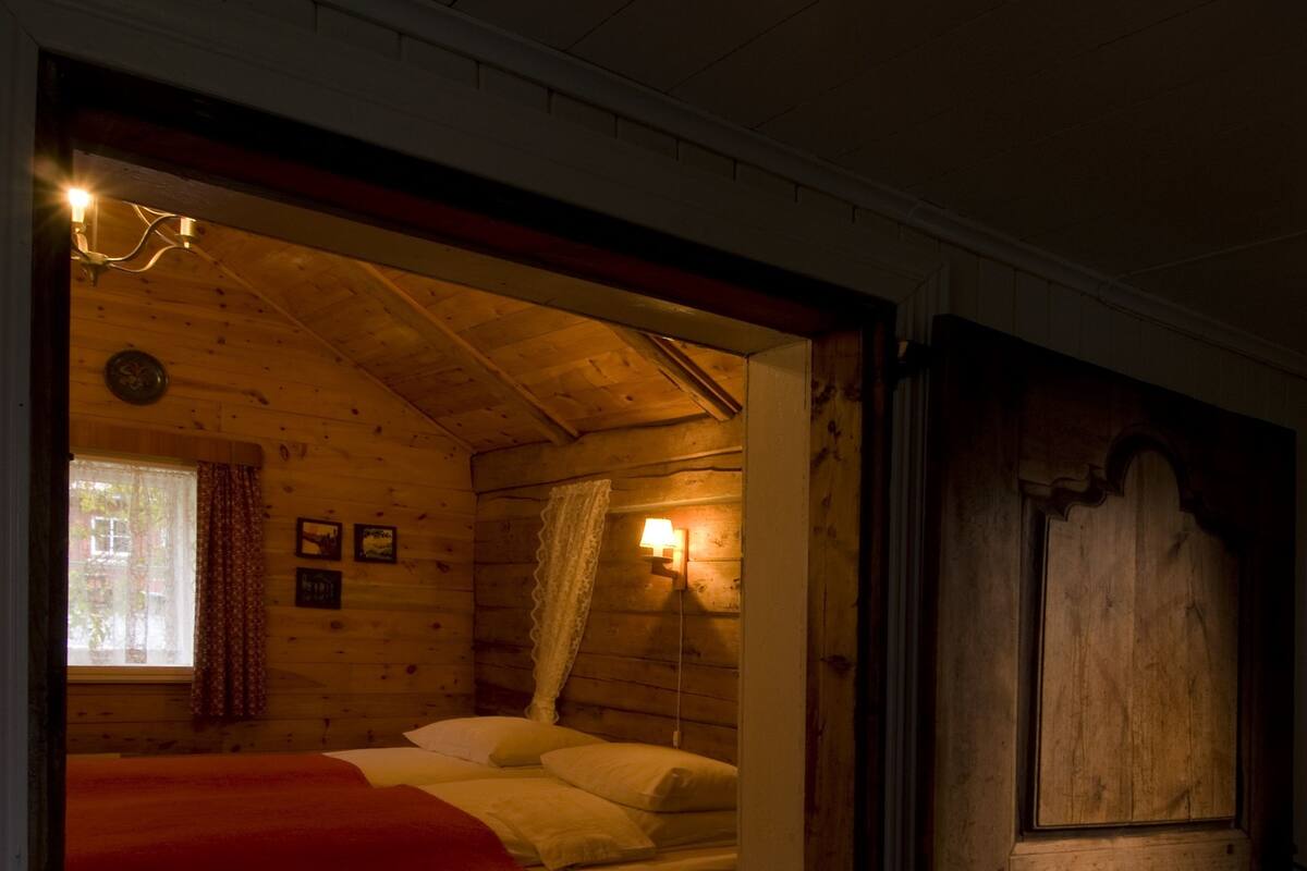Una habitación de cabaña de madera con una cama, una ventana con cortinas y una lámpara fijada a la pared.