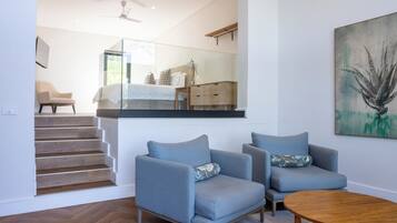Una sala de estar moderna con piso de madera en patrón de espiga, dos sillones azules, una mesa de centro de madera y una escalera de cristal que lleva a un nivel superior.