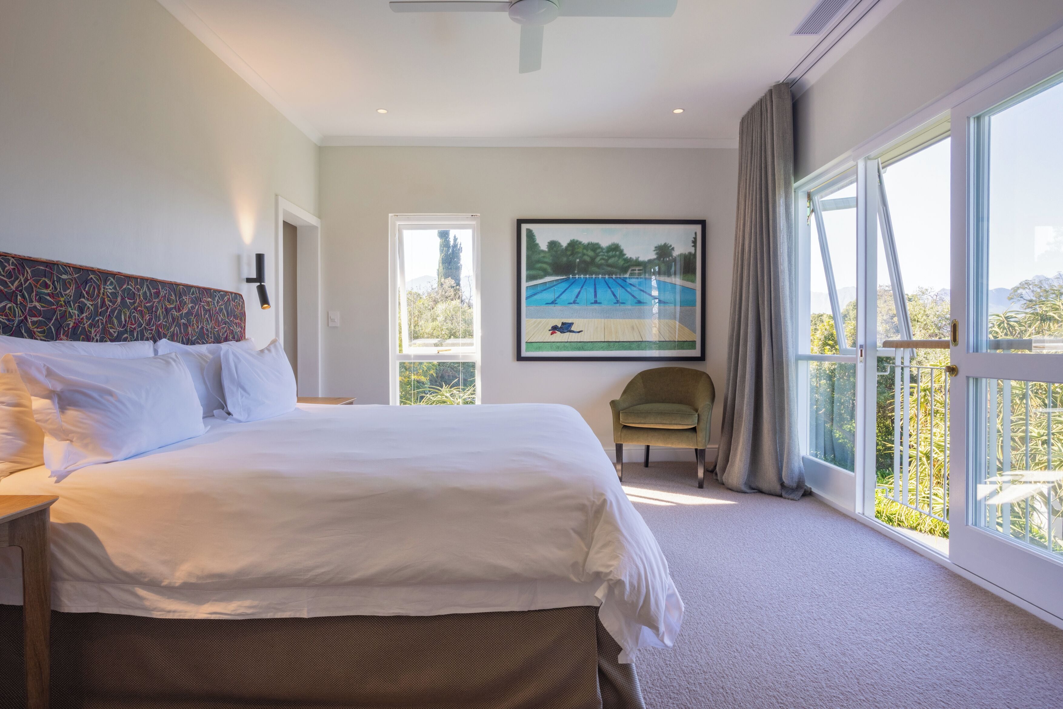 A hotel room with a large bed, a chair, a painting, and a view of a swimming pool.