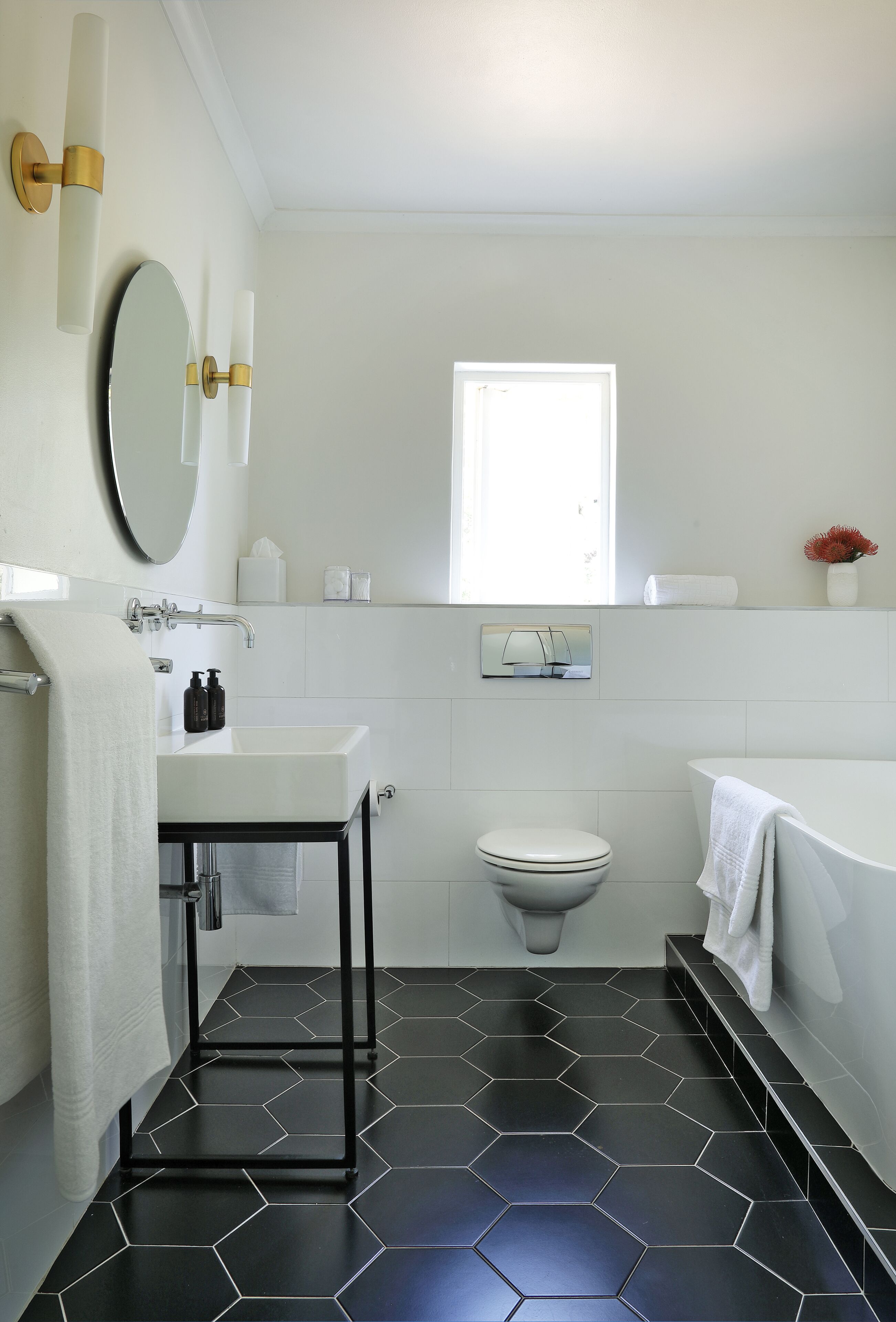 A modern bathroom with a white sink, toilet, and bathtub, black hexagonal tiles, and a window providing natural light.