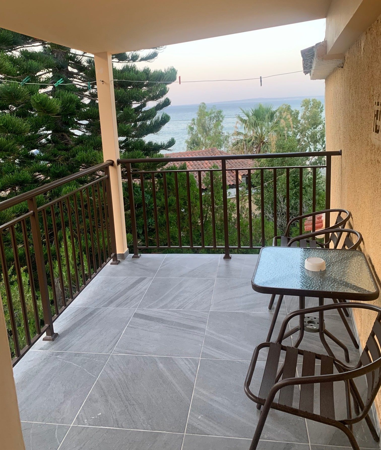 Een balkon met een glazen tafel en stoelen, een betegelde vloer en uitzicht op zee en bomen.