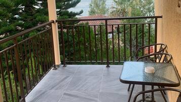 Een balkon met een glazen tafel en stoelen, een betegelde vloer en uitzicht op zee en bomen.