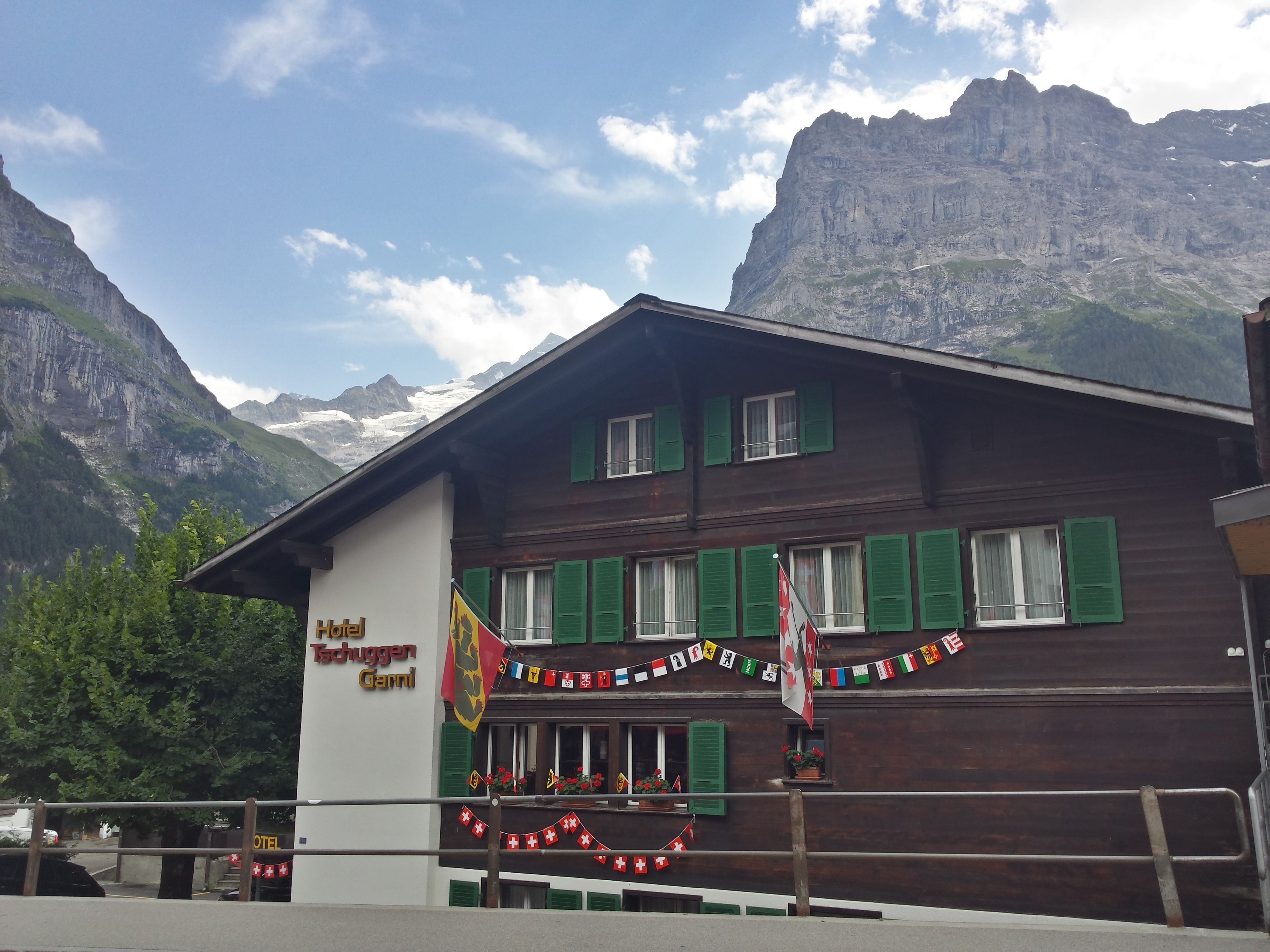 A hotel with a mountain backdrop, featuring a wooden facade, green shutters, and a sign indicating it is a hotel.