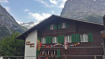 A hotel with a mountain backdrop, featuring a wooden facade, green shutters, and a sign indicating it is a hotel.