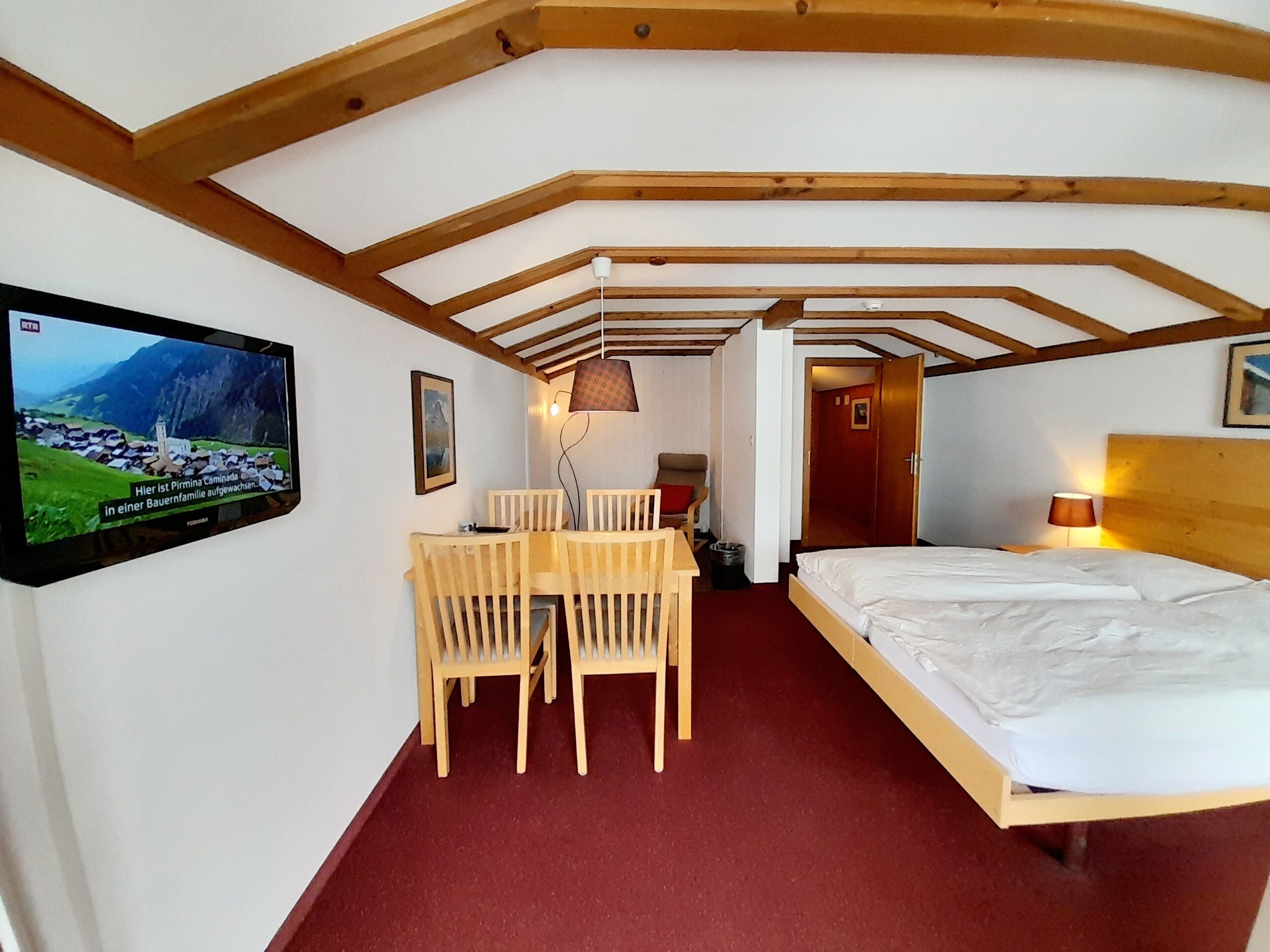 Una habitación de hotel con una cama, un televisor montado en la pared, una mesa de madera con sillas y una alfombra roja.