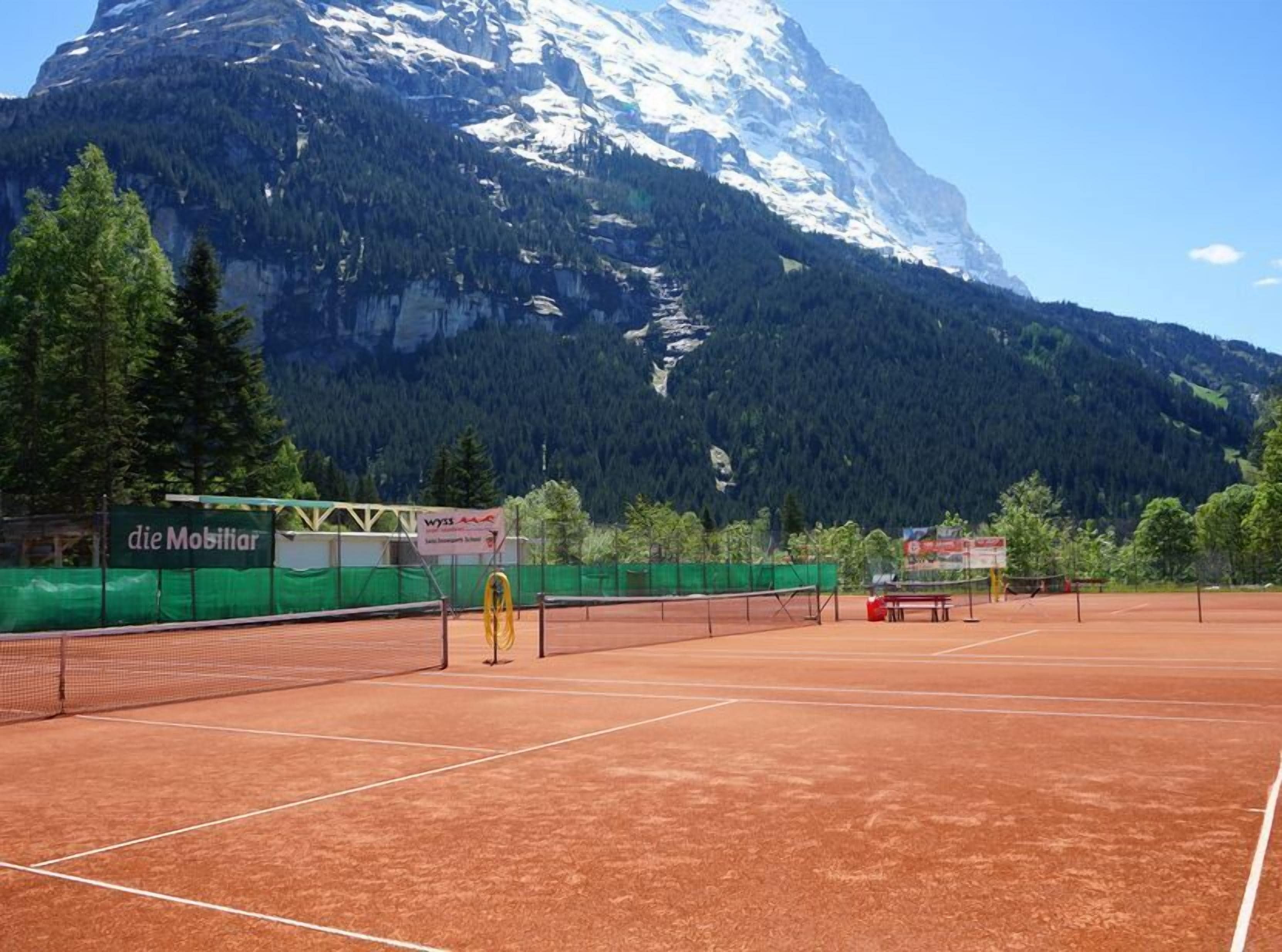 Una cancha de tenis al aire libre con montañas al fondo, rodeada de árboles verdes y un cartel de 'die Mobiliar'.