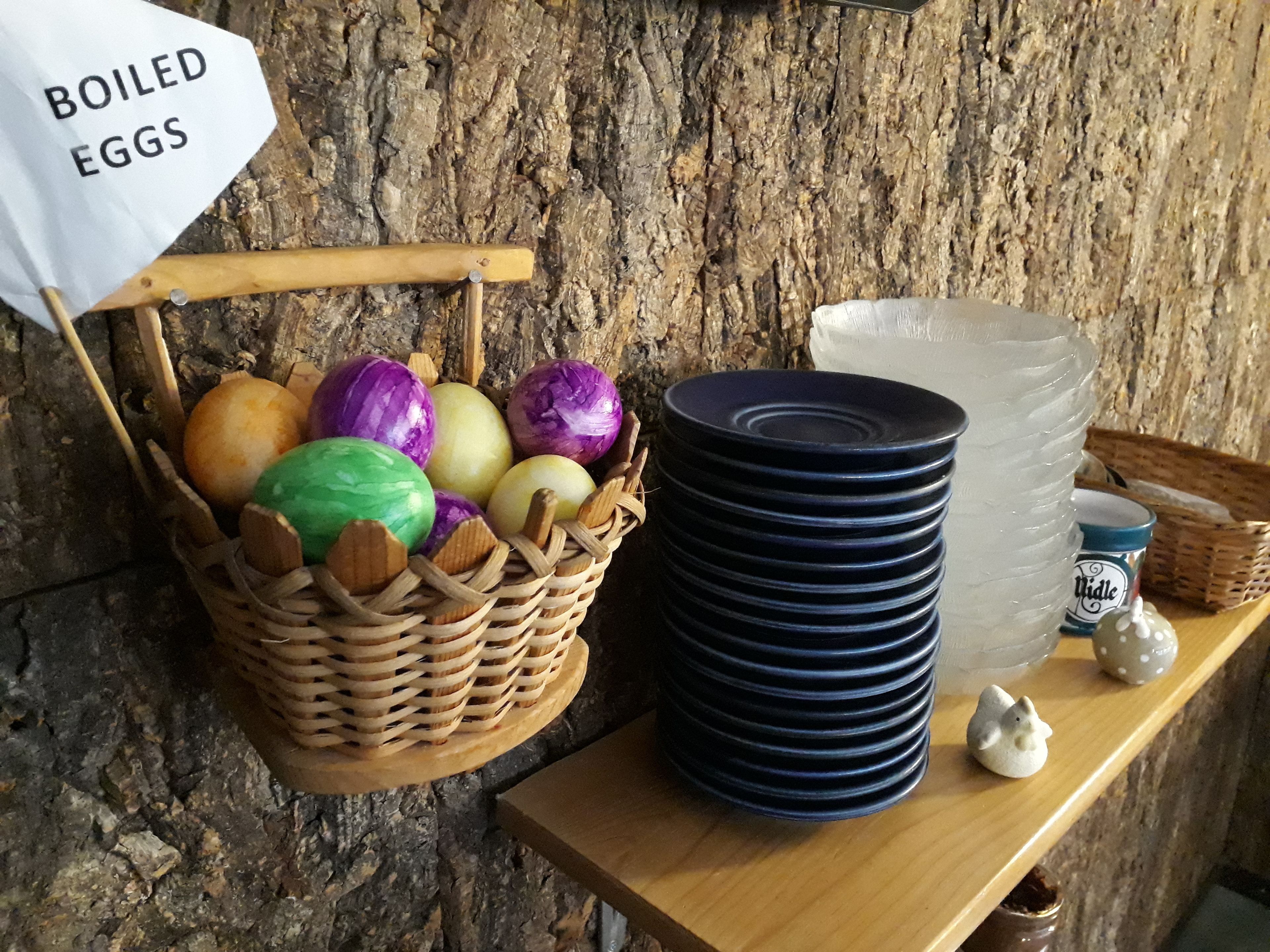 Una cesta de madera llena de huevos cocidos de colores, una pila de platos azules y un recipiente de vidrio grande sobre una estante de madera.