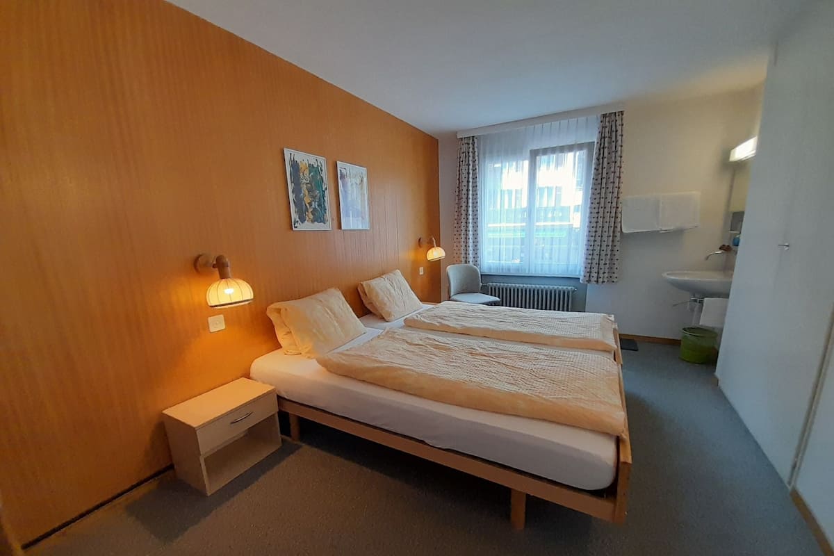 Habitación de hotel con pared de madera, cama con dos almohadas, mesita de noche, silla, ventana con cortinas, radiador, lavamanos y un armario.