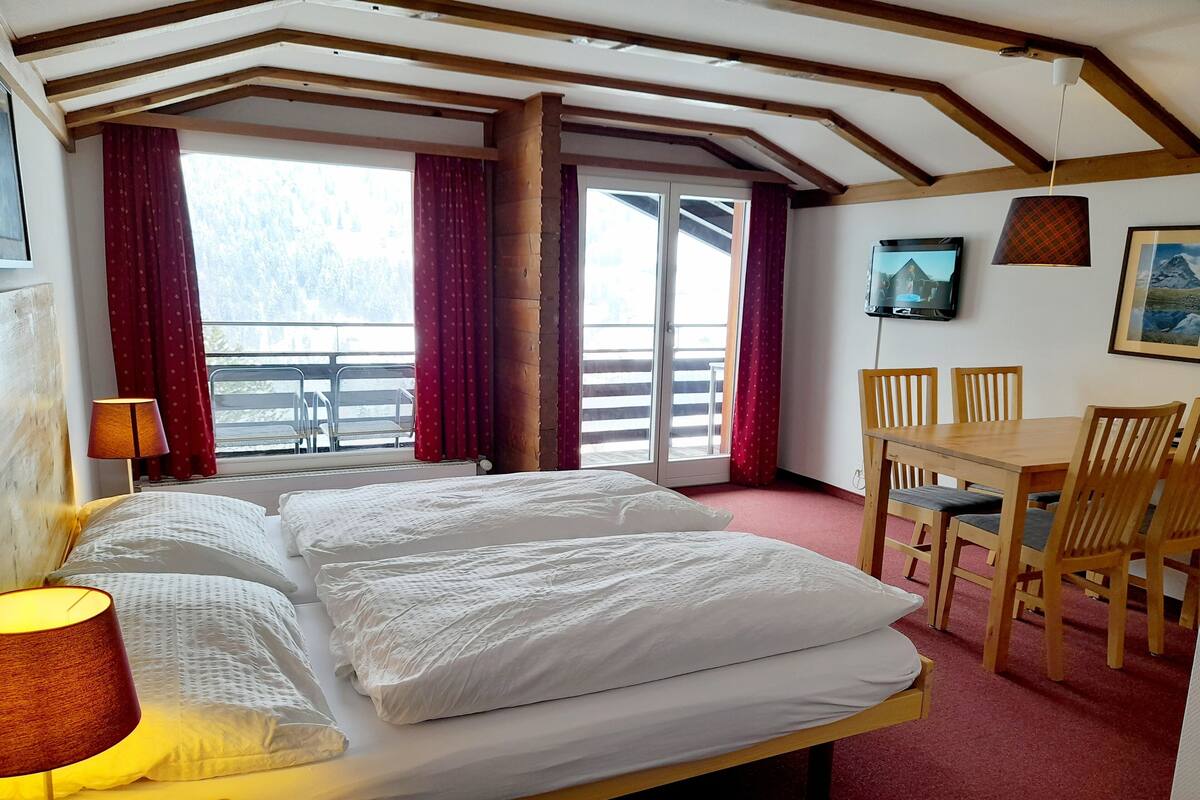 Habitación de hotel con una cama grande, una mesa de madera con sillas, un televisor en la pared y un balcón con cortinas rojas.