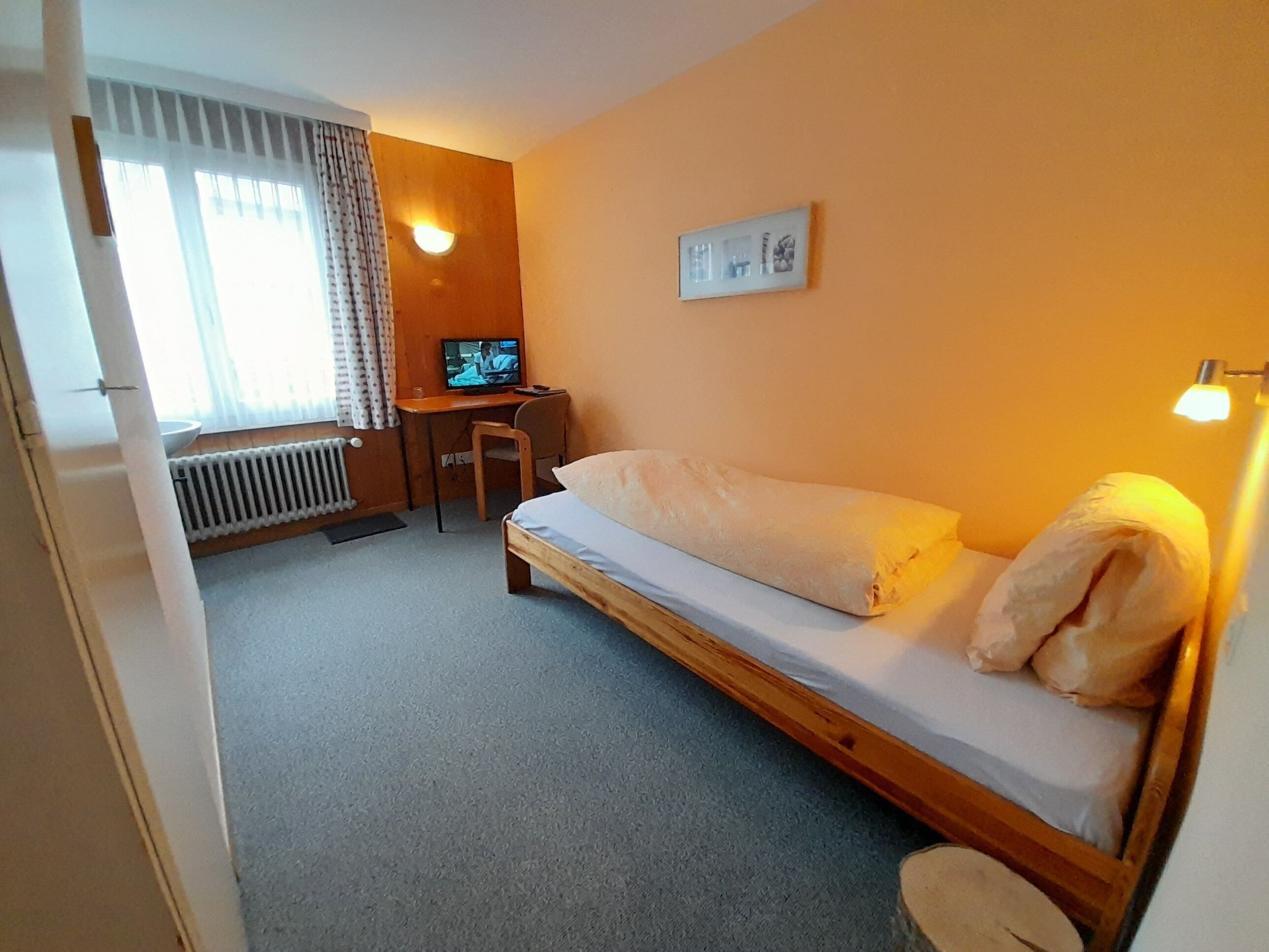 Une petite pièce comprenant un lit, un bureau, une chaise, un téléviseur et un radiateur.