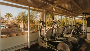 A gym with treadmills and weight machines, large windows, and a view of palm trees and buildings. - Maspalomas Resort by Dunas (San Bartolomé de Tirajana)