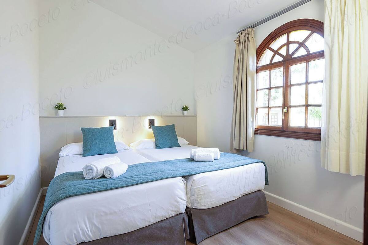 A double bed with white and blue bedding, two bedside lamps, a window with curtains, and a small plant on the nightstand.