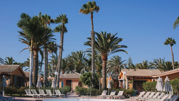 A pool area with lounge chairs and umbrellas, surrounded by palm trees and buildings with tiled roofs. - Maspalomas Resort by Dunas (San Bartolomé de Tirajana)