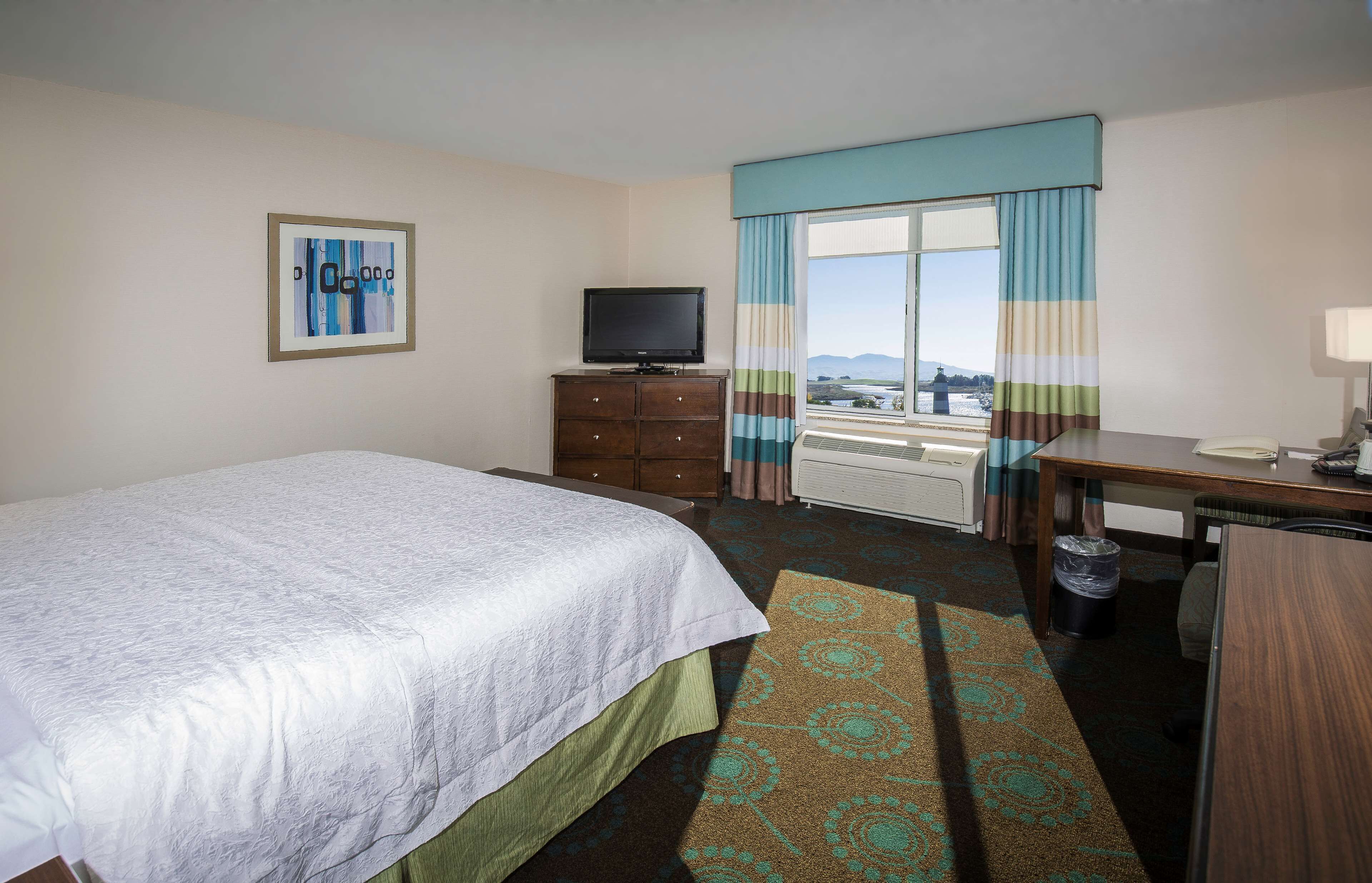 Habitación de hotel con cama, escritorio, televisor, ventana con cortinas y vista a la ciudad.