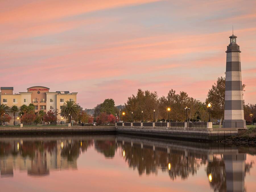 A waterfront view with a lighthouse, a building with a sign, and a calm river reflecting the sky.