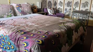 Une chambre d’hôtel avec un grand lit, un fauteuil violet, une petite table et des œuvres d’art traditionnelles accrochées aux murs.