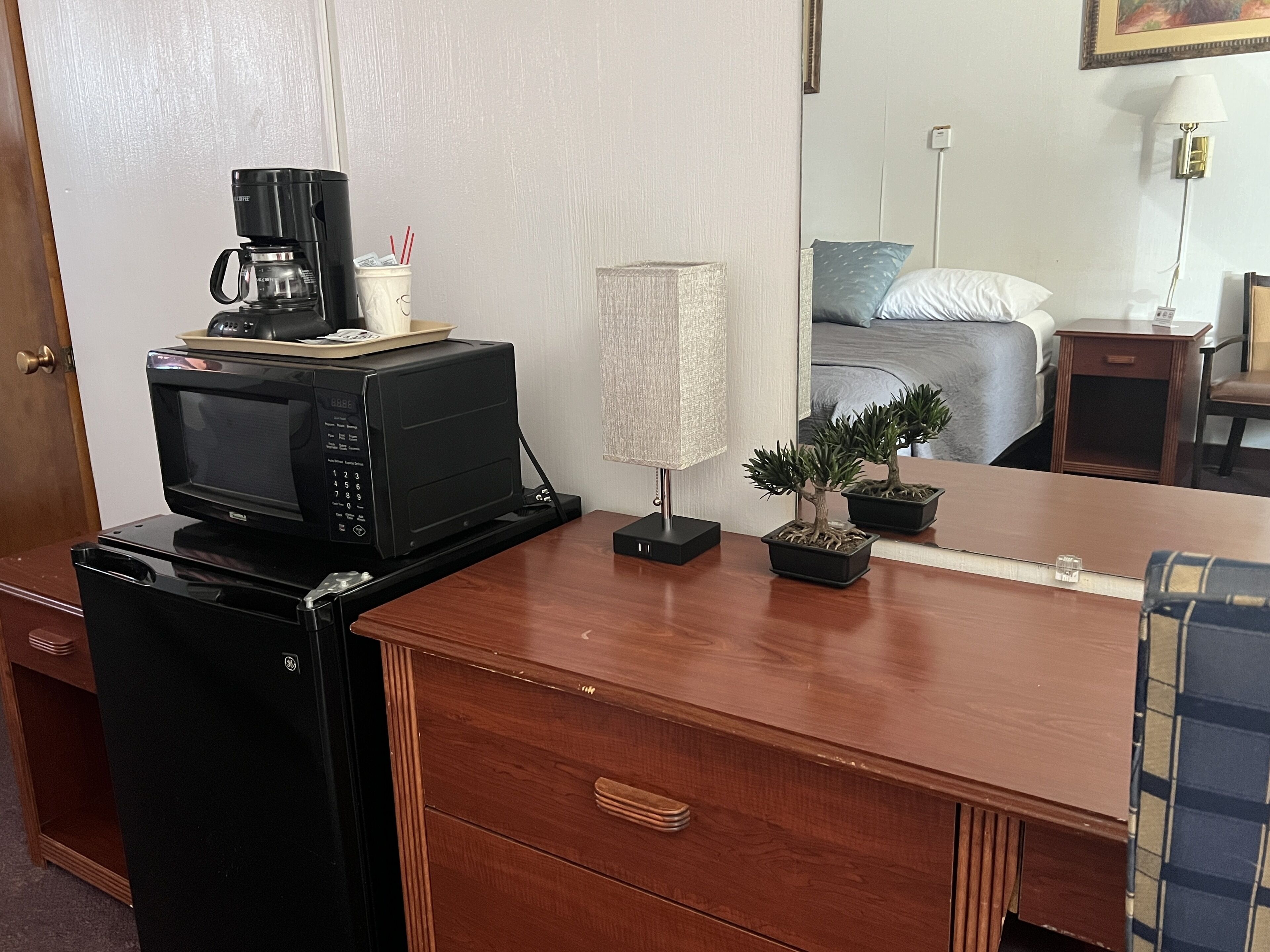 Een compacte hotelkamer met een houten bureau, een zwarte koelkast, een magnetron, een koffiezetapparaat, een lamp en een kleine potplant.