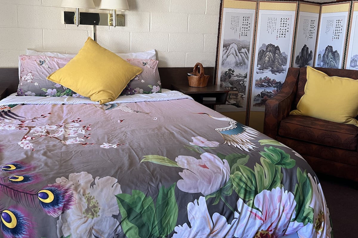 A neatly made bed with floral bedding, a pair of lamps, and a traditional Chinese screen with paintings.