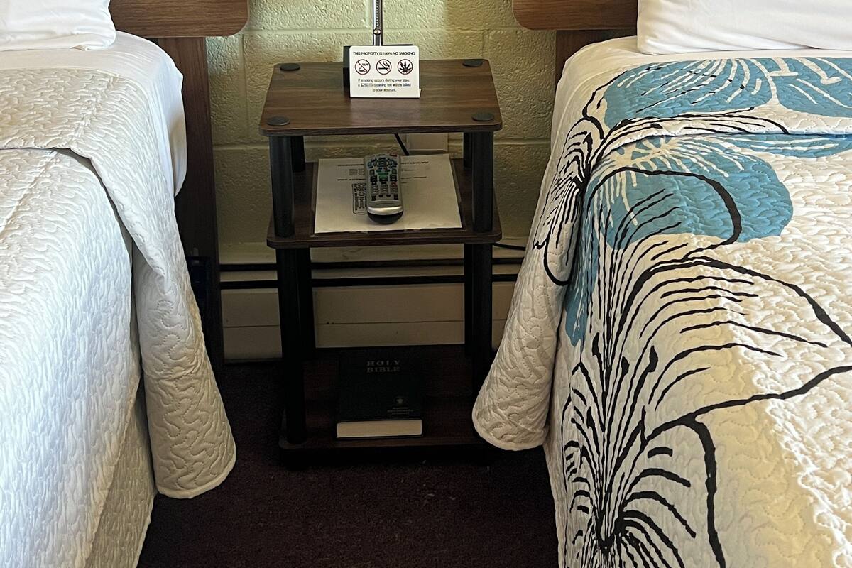 A hotel room with two beds, a nightstand with a lamp, a remote control, and a Bible.