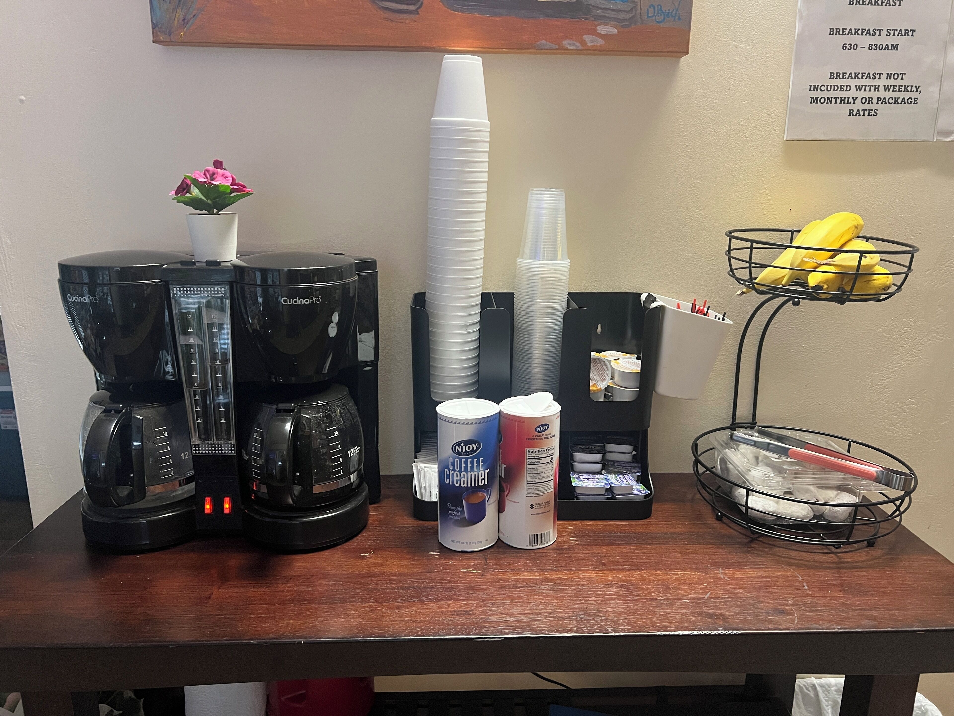 Una estación de café con dos cafeteras, una pila de vasos desechables, un recipiente con leche para café y una cesta de frutas.