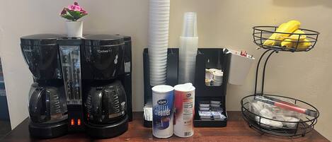 Una estación de café con dos cafeteras, una pila de vasos desechables, un recipiente con leche para café y una cesta de frutas.