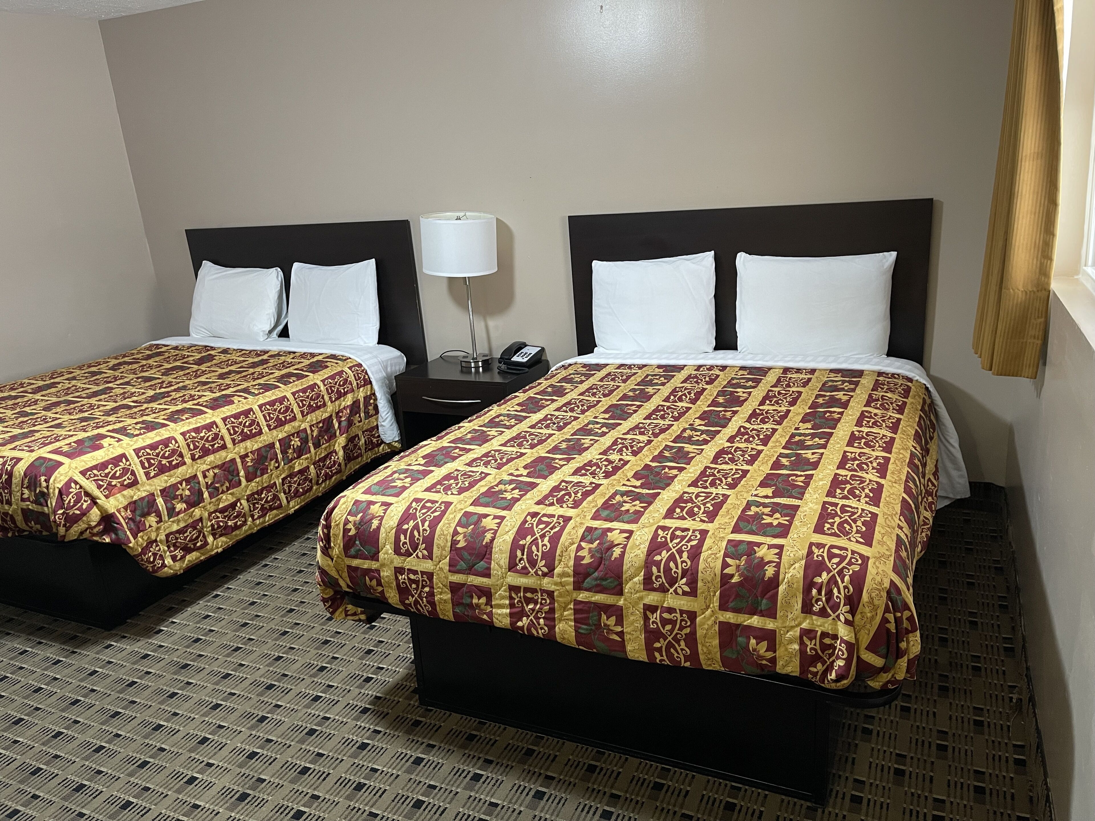 A hotel room with two beds, each with a patterned bedspread, a bedside table with a lamp, and a plain wall.
