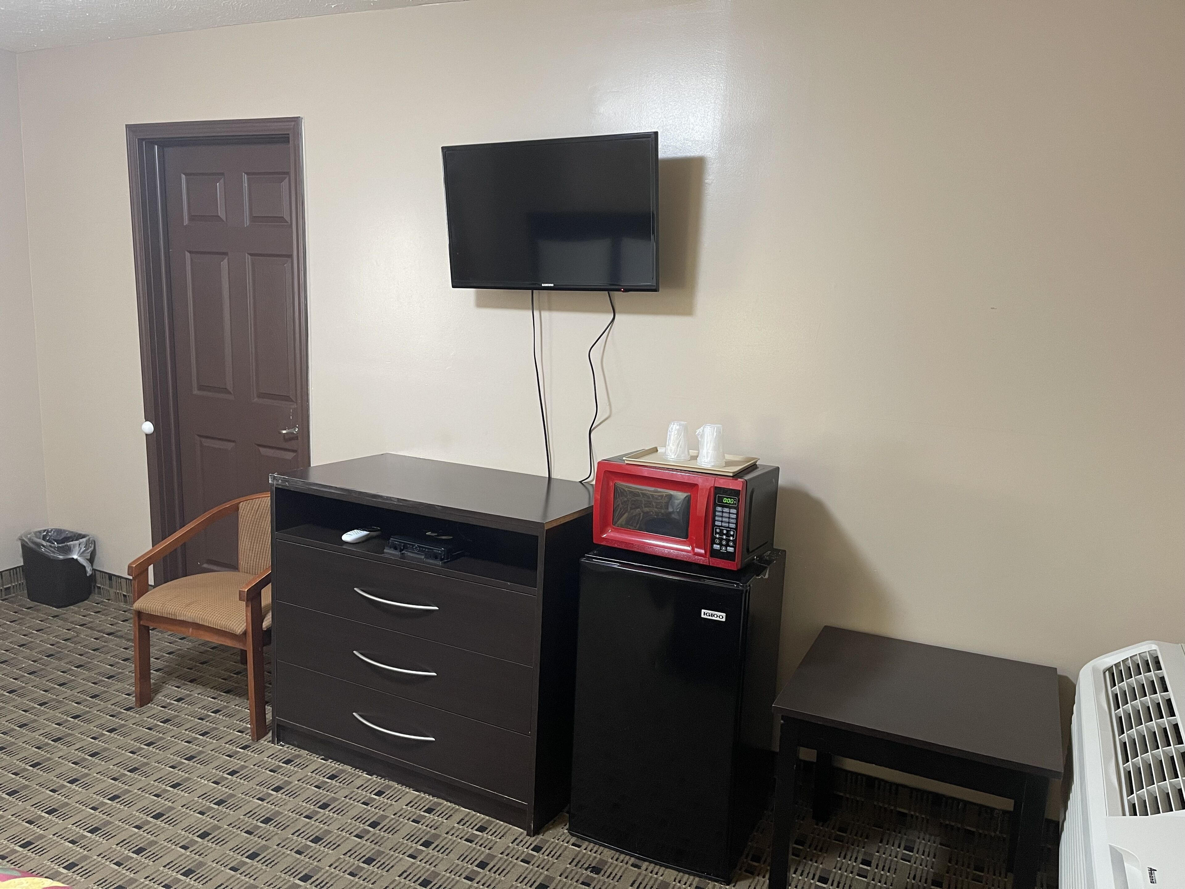 A compact room with a desk, a chair, a microwave, a television, and a small table.