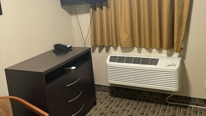 A hotel room with a dark wooden desk, a flat-screen TV mounted on the wall, a window with beige curtains, and a white air conditioning unit. - Economy Inn And Suites Ashtabula (Ashtabula)