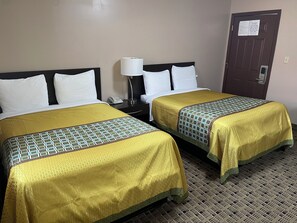 Dos camas dobles con colchas estampadas en amarillo y verde, una mesita de noche con lámpara, y una puerta con señal de seguridad contra incendios.
