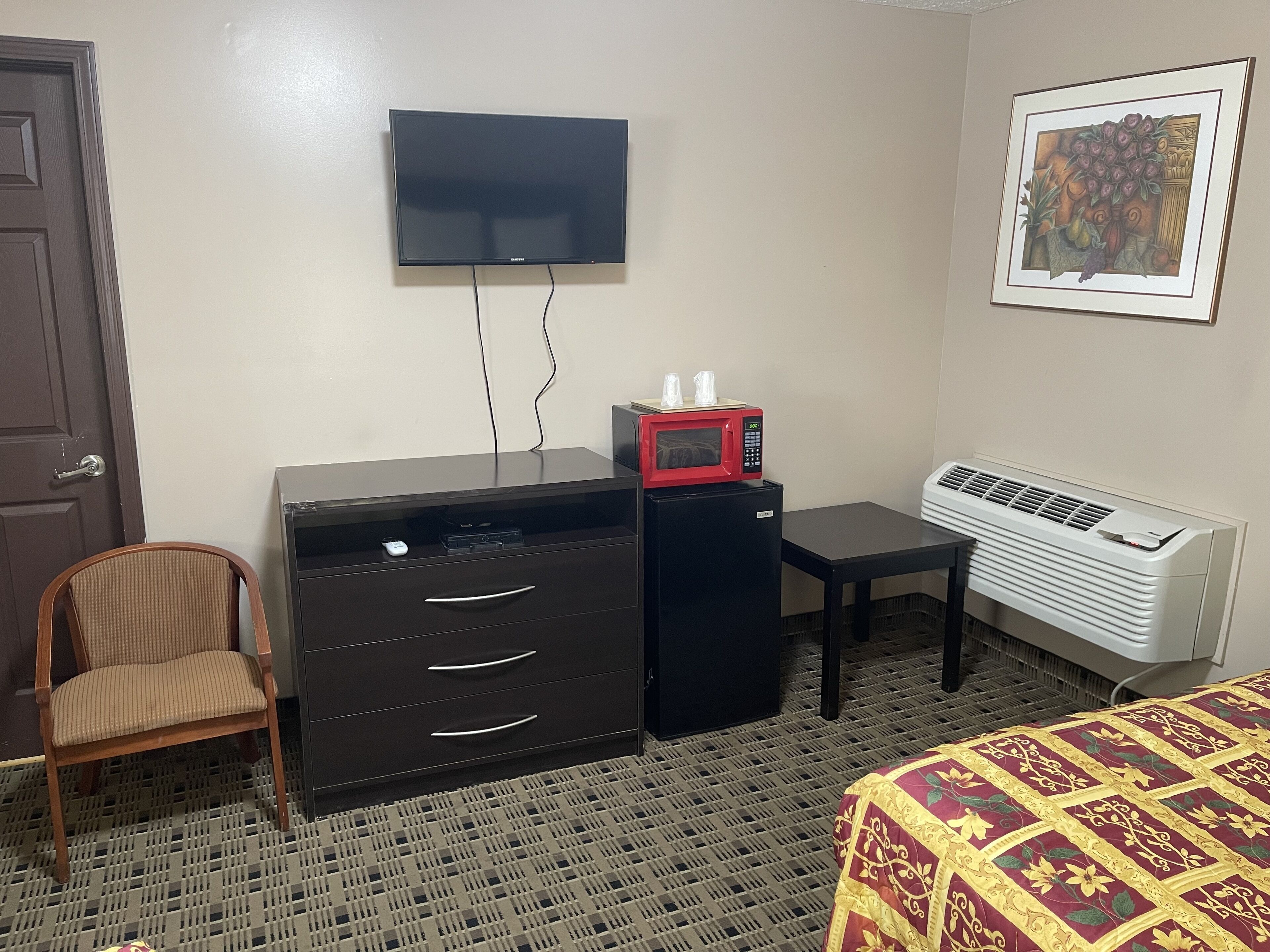 Habitación de hotel con televisión de pantalla plana en la pared, un refrigerador pequeño negro con un microondas encima, una silla, un escritorio con cajones y un radiador.