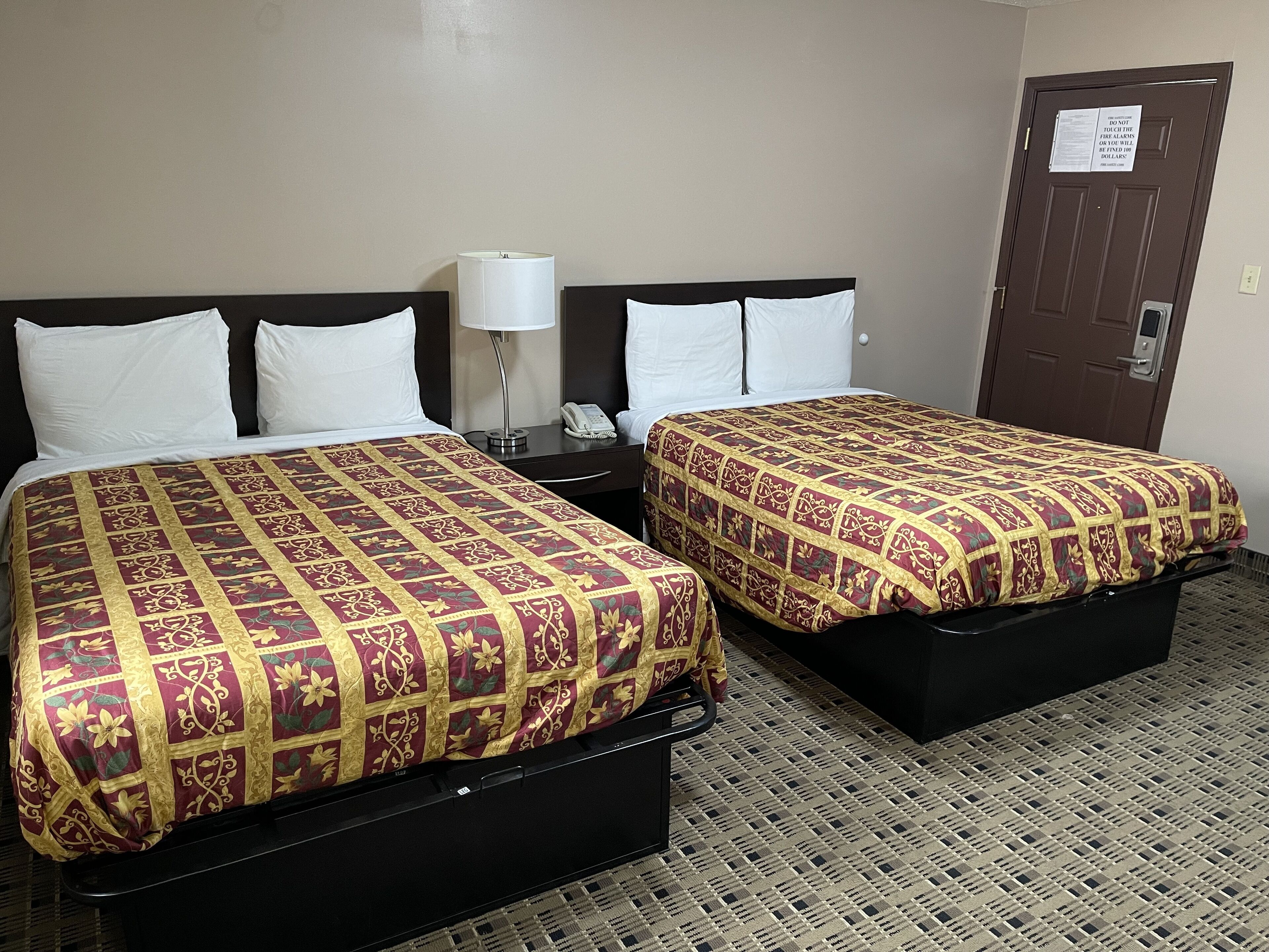 A hotel room with two beds, each with a patterned bedspread, a bedside table with a lamp, and a door with a security notice.