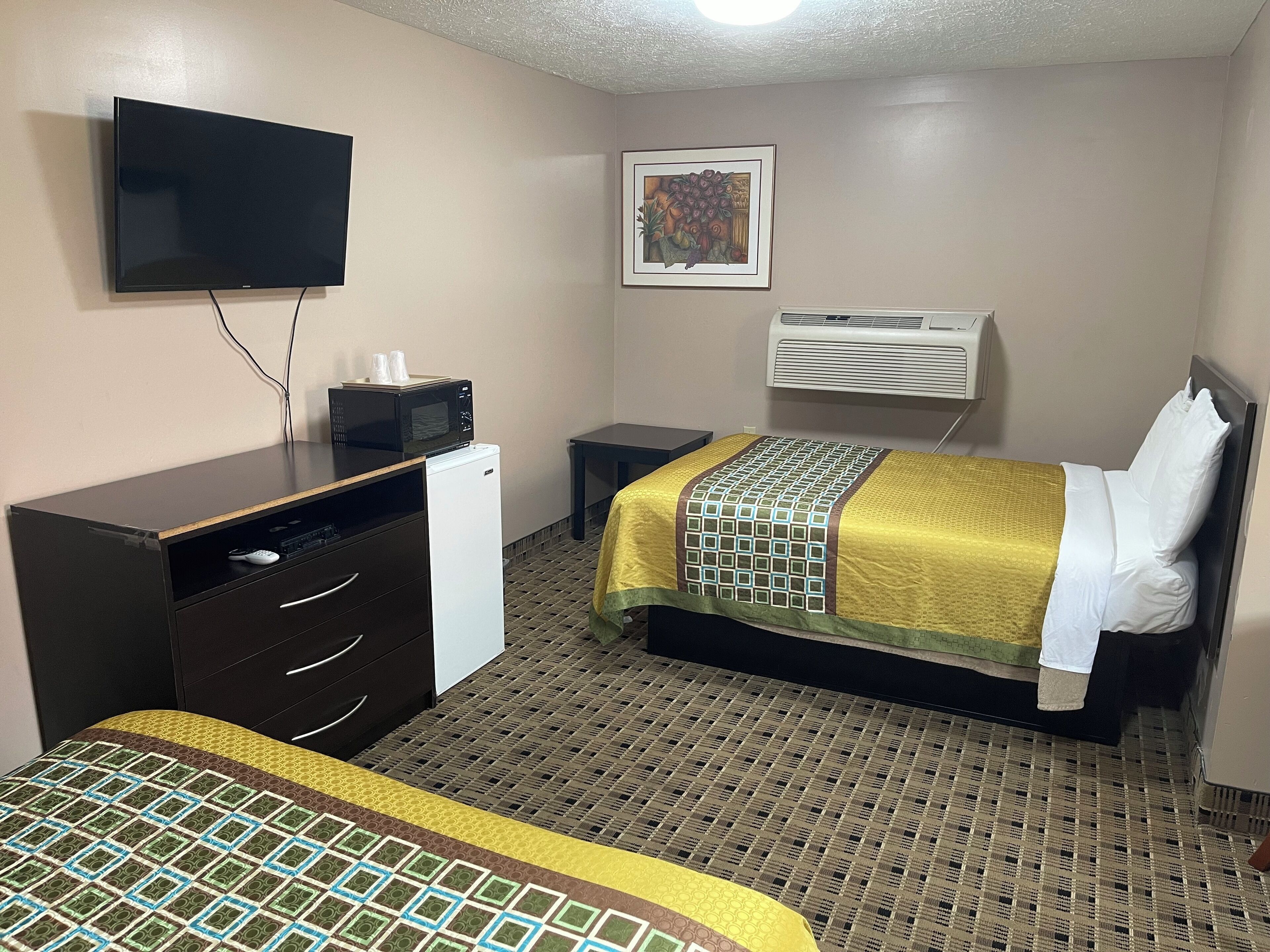 A hotel room with a bed, a desk with a computer, a TV, an air conditioner, and a framed picture on the wall.