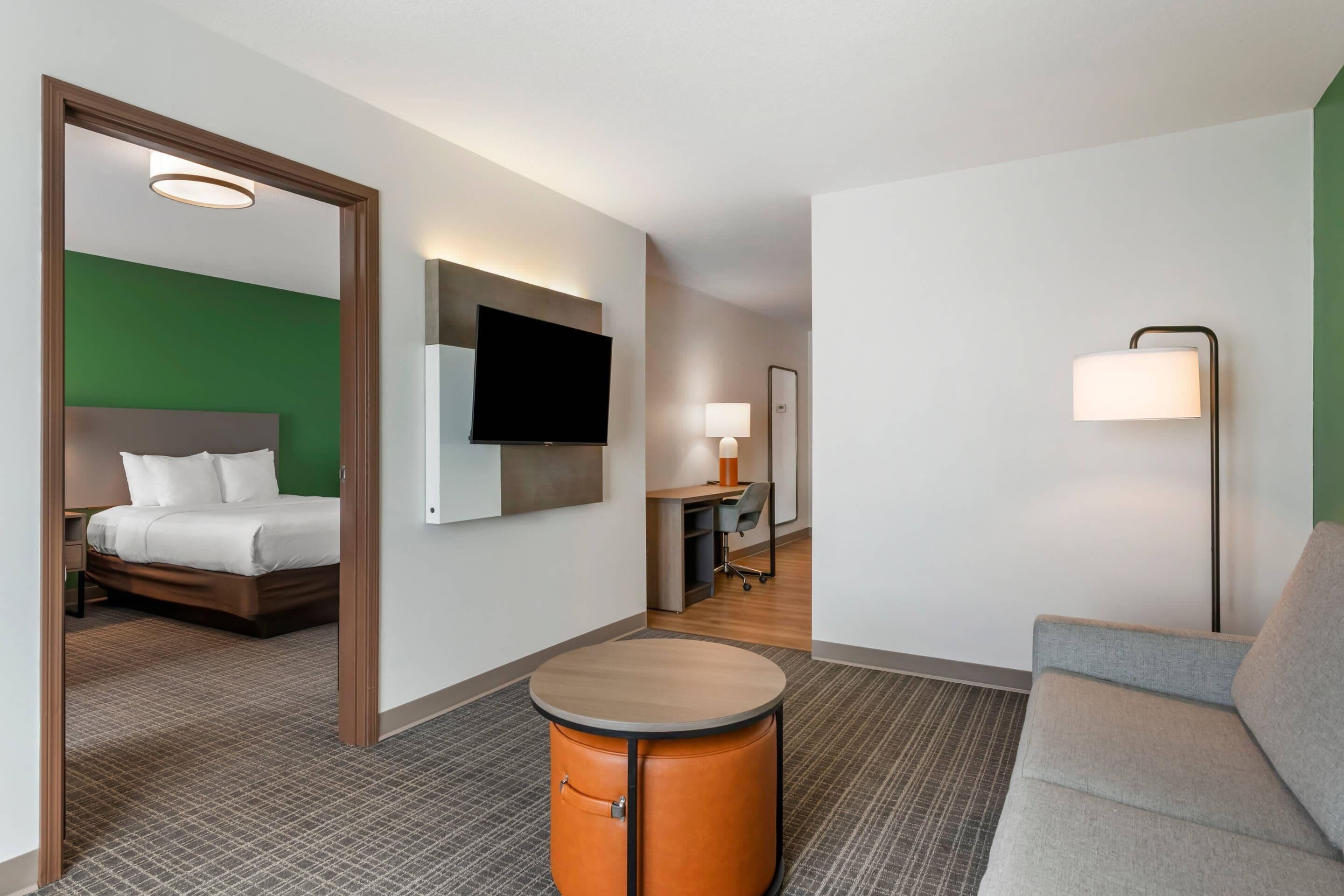 Une chambre d’hôtel comprenant un lit, une télévision, un bureau avec une chaise, un pouf rond orange et un canapé gris.