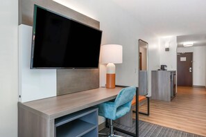 A hotel room with a flat-screen TV, a desk with a lamp, a blue chair, and a mirror.